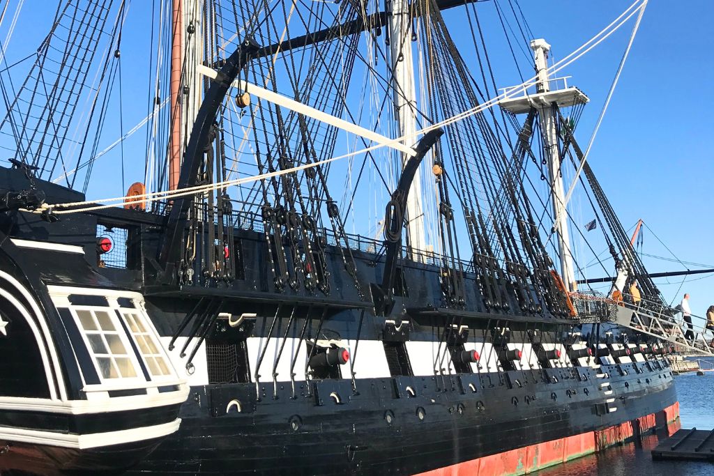 Side view of the USS Constitution, a historic naval warship docked in Boston, also known as 'Old Ironsides.' The ship's black and white hull, tall masts, and intricate rigging are prominently featured under a clear blue sky.