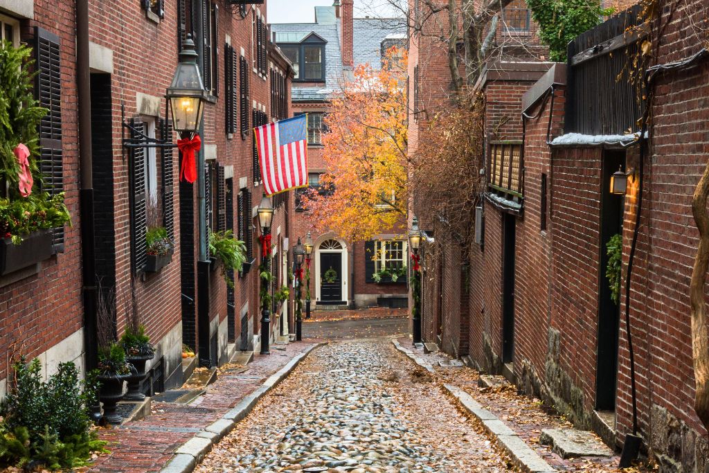 View of Acorn Street in Boston's historic Beacon Hill neighborhood, featuring cobblestone pavement lined with brick townhouses decorated with American flags and holiday wreaths. The narrow street is framed by gas lanterns and fall foliage, capturing a charming autumn scene.