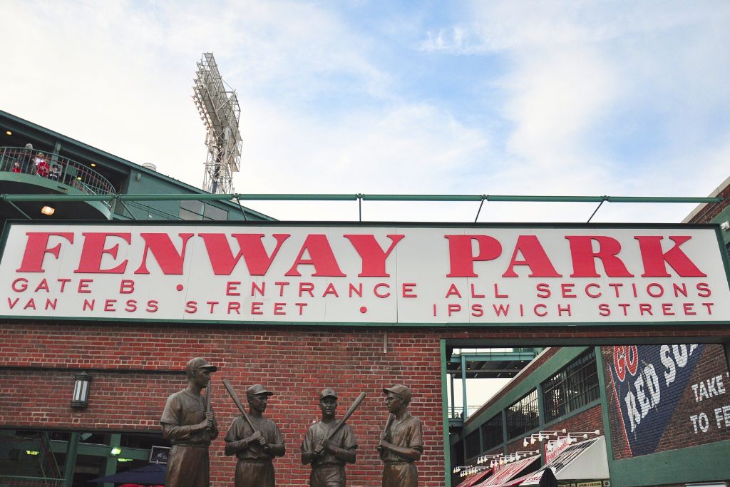 Fenway Park entrance sign above Gate B, displaying 'Fenway Park' in large red letters along with directions to Van Ness and Ipswich Streets. Below the sign are statues of legendary baseball players, capturing the historic atmosphere of the iconic ballpark in Boston.