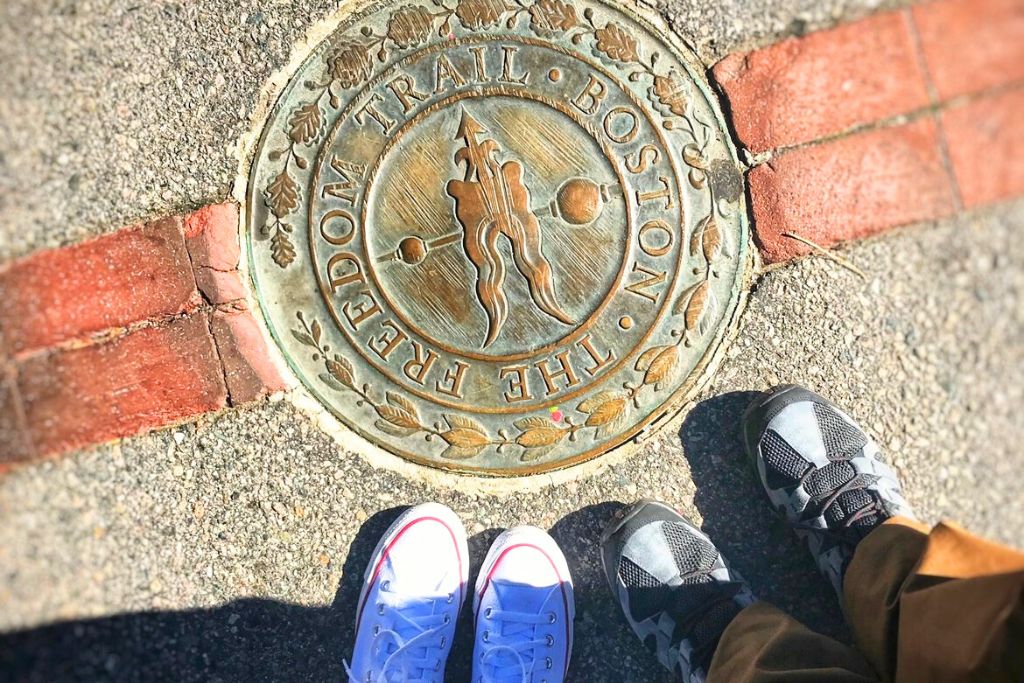 Bronze marker for Boston's Freedom Trail embedded in the sidewalk, with two pairs of feet visible, one in white sneakers and the other in gray, standing on either side of the brick-lined path. The marker reads 'The Freedom Trail Boston,' is one of the most popular Things to do in Boston in October.