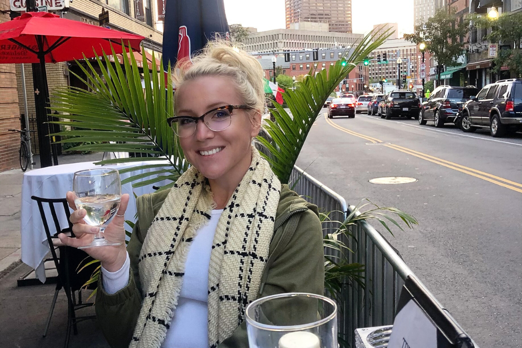 Kate, smiling while holding a glass of wine, seated at an outdoor restaurant during a food tour in Boston. The background shows a city street with cars, Italian flags, and restaurants, creating a lively urban atmosphere.