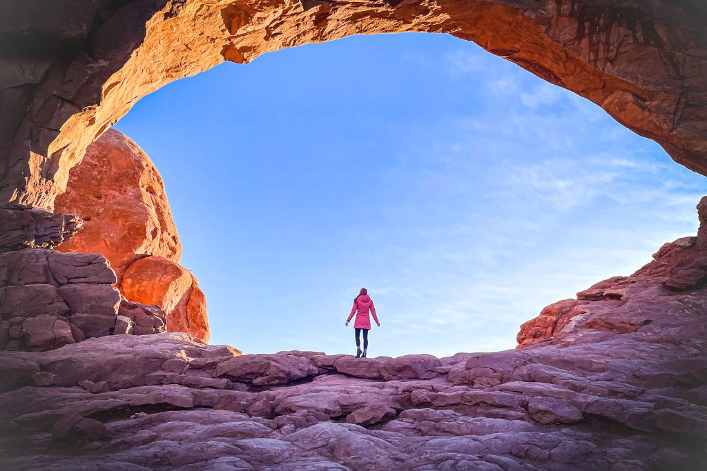 Kate is standing under a natural rock arch at sunrise, gazing at the expansive blue sky in a moment of reflection, symbolizing the concept of transformative travel and personal growth through exploring nature.