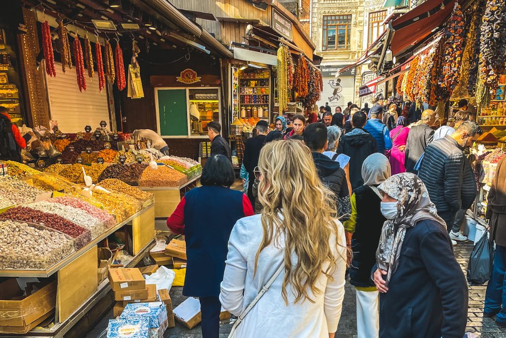 Kate walks through the bustling spice market in Istanbul, Turkey, surrounded by stalls filled with colorful spices, dried fruits, and other goods. The vibrant market is packed with locals and tourists, creating a lively atmosphere as she navigates the narrow streets of the large city. The scene captures the rich culture and sensory experience of exploring Istanbul.