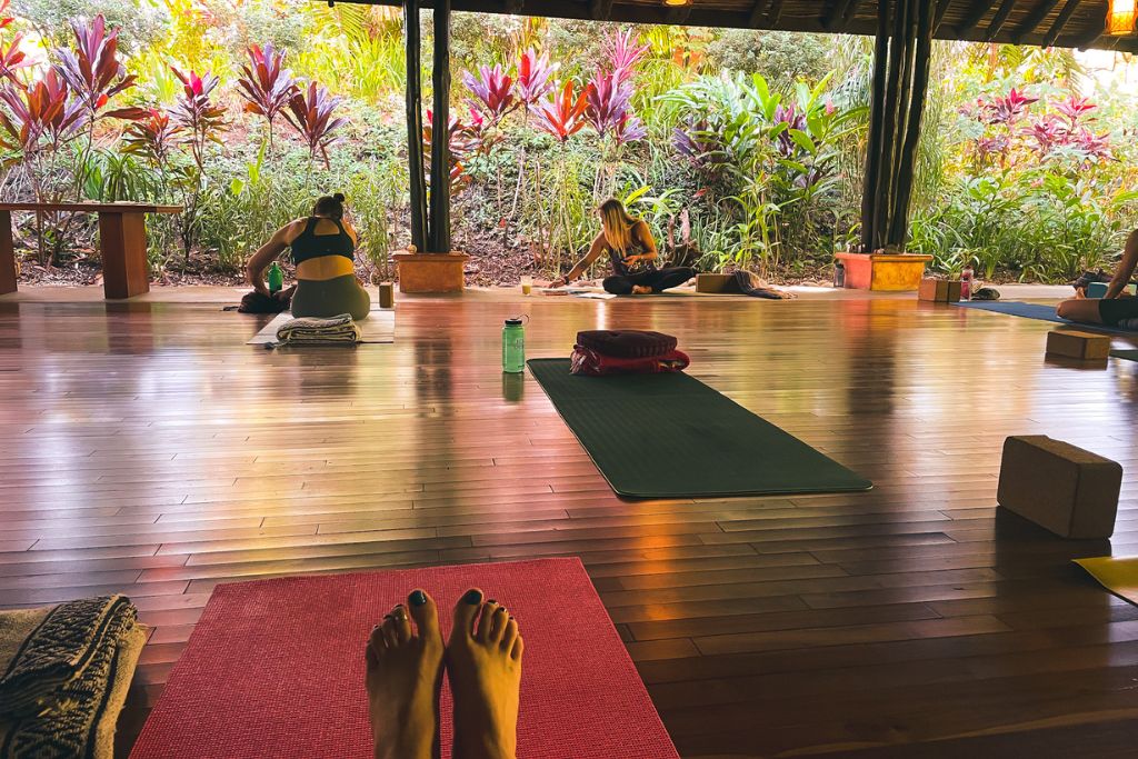 Participants are gathered in a serene, open-air yoga studio surrounded by lush tropical plants during a yoga retreat in Costa Rica. Yoga mats are spread out on the polished wooden floor, with some individuals preparing for their session while others sit quietly in reflection. The peaceful, nature-filled setting provides a calming environment for mindfulness and relaxation.