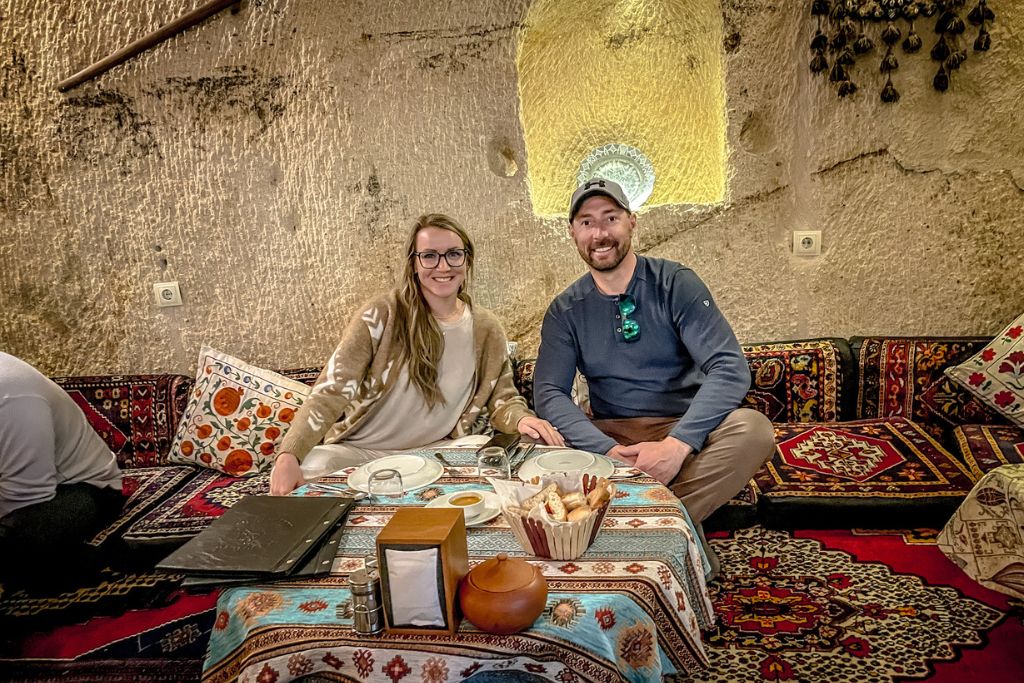 Kate and her husband sit at a low table on the ground inside a cozy cave restaurant in Cappadocia, Turkey, enjoying a traditional meal. They are surrounded by vibrant patterned cushions and rugs, adding to the authentic atmosphere of the unique dining experience. The setting highlights the cultural richness and distinctive architecture of the region.