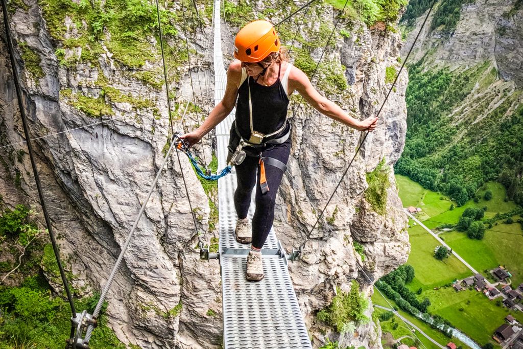 Kate carefully walks across a narrow suspension bridge high above a valley during an adventure in Mürren, Switzerland. She wears a helmet and harness for safety, gripping the cables while navigating the bridge, with the dramatic cliffs and lush green landscape far below. The scene captures both the thrill and beauty of the alpine adventure.