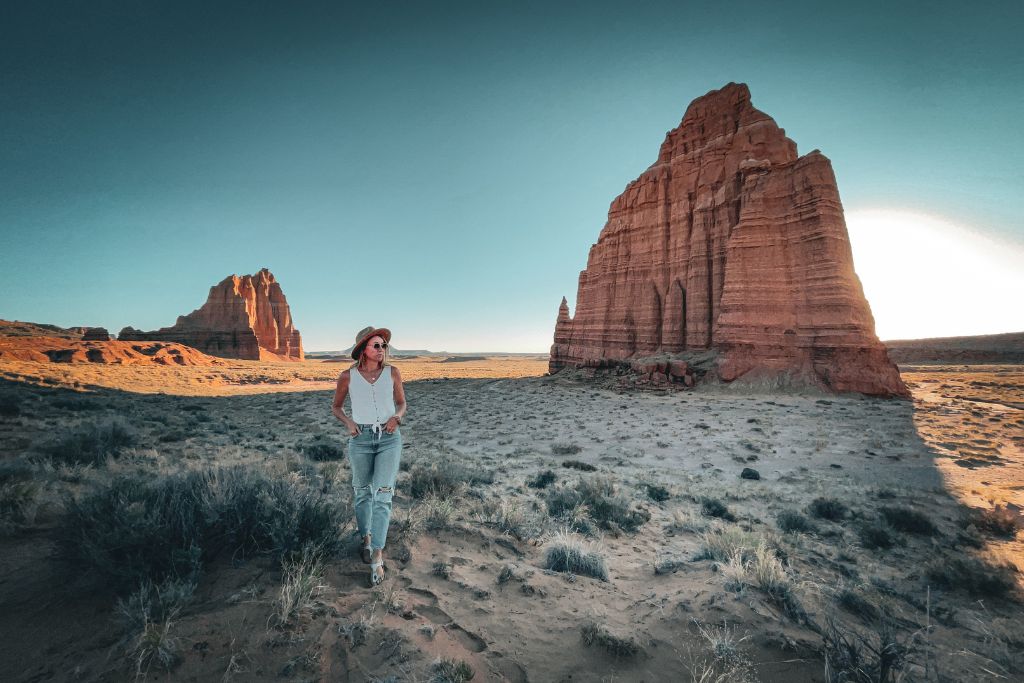 Kate walks through the desert landscape in Utah, with the towering rock formations of the Temple of the Sun and the Moon behind her. She is dressed casually in a white tank top, jeans, and a wide-brimmed hat, capturing the beauty of the rugged terrain as the sunlight casts dramatic shadows on the sandstone formations. The scene evokes a sense of awe and adventure in this iconic natural setting.