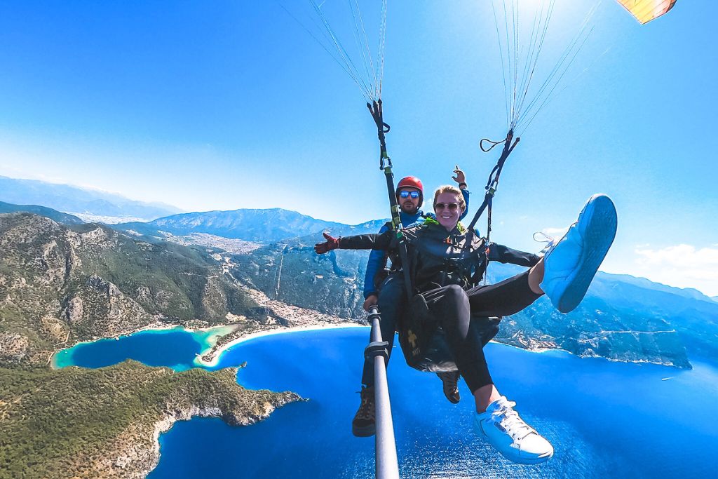 Kate is paragliding for the first time in Oludeniz, Turkey, soaring high above the stunning turquoise coastline and lush green mountains. She smiles excitedly, legs outstretched, as she and her guide enjoy the breathtaking views of the landscape below. The clear blue sky and vast scenery highlight the thrill and beauty of this adventurous experience.