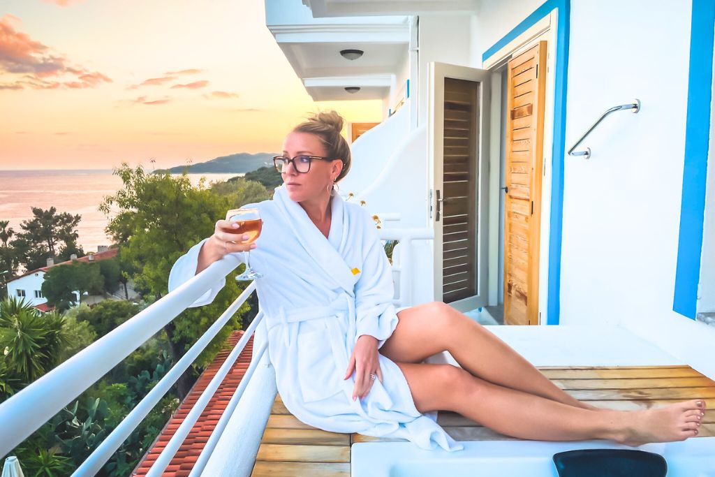 Kate sits on her hotel balcony overlooking the Mediterranean Sea, wearing a white robe and holding a glass of wine as she watches the sunset. The soft pastel colors of the sky blend with the peaceful ocean view, creating a serene and relaxing atmosphere. This moment captures the tranquility of unwinding in a beautiful seaside location.