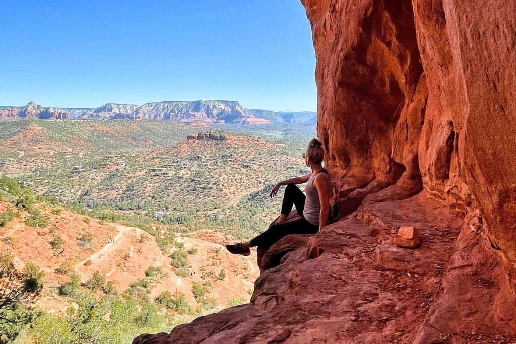 Kate sits on a ledge inside a red rock cave, gazing out at the expansive and spiritual landscape of Sedona, Arizona. The vibrant red rock formations and lush valleys stretch out under a clear blue sky, creating a moment of peaceful reflection. The scene captures the tranquil and mystical energy of this renowned spiritual destination.