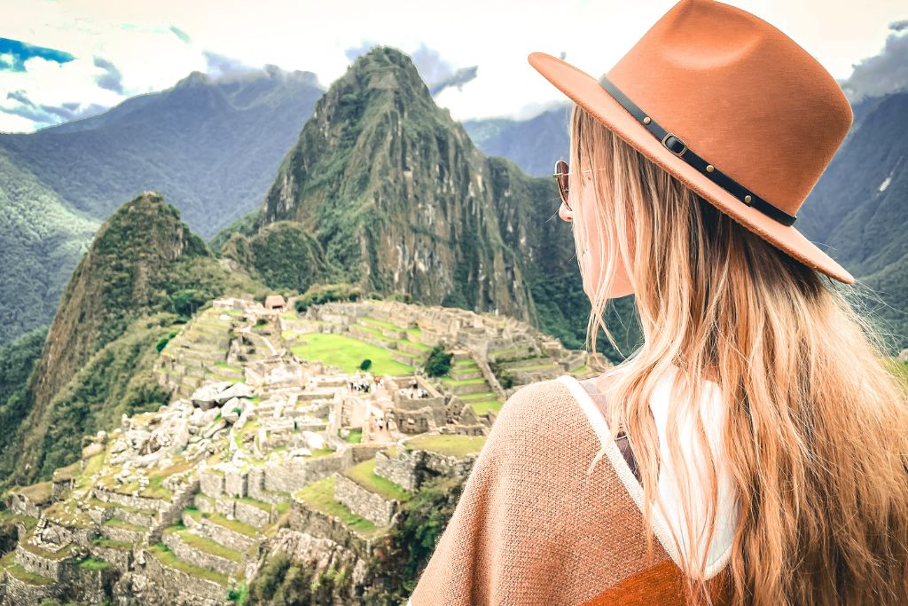 Kate stands overlooking the ancient Incan city of Machu Picchu in Peru, wearing a tan hat and gazing at the majestic mountain backdrop. The ruins of the stone terraces and buildings stretch out below, framed by the lush green Andes mountains. The scene captures the awe-inspiring beauty and historical significance of this iconic site.