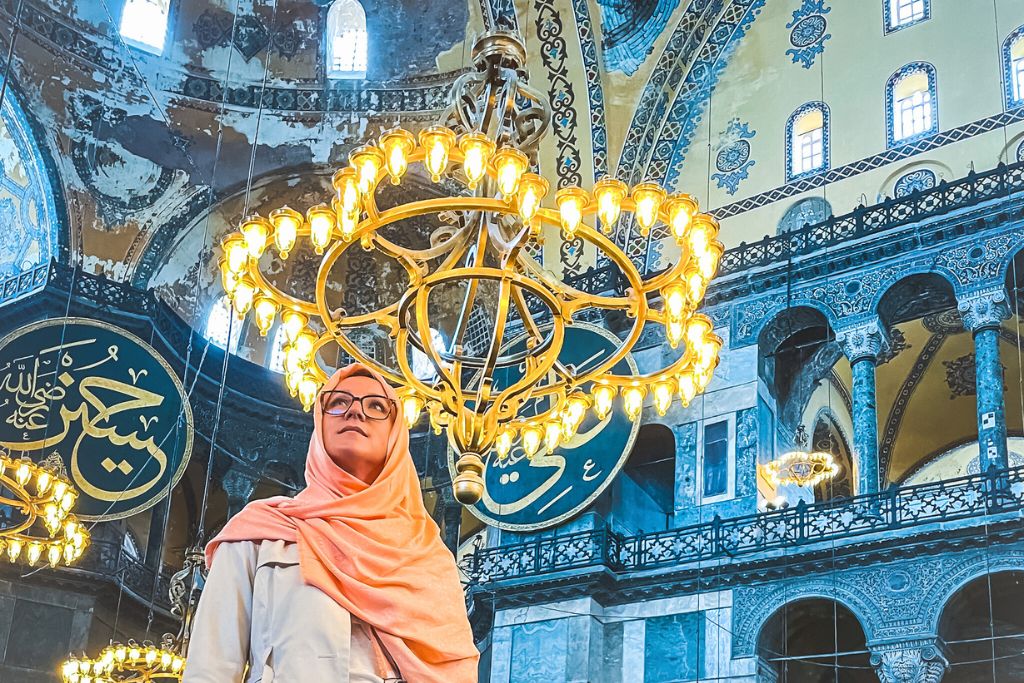 Kate is wearing a pink headscarf and stands gazing up at the intricate architecture inside the Hagia Sophia Mosque in Istanbul, Turkey. A large illuminated chandelier hangs from above and Arabic calligraphy and ornate designs decorate the historic Mosque's walls. The scene captures the grandeur and serenity of the sacred space.