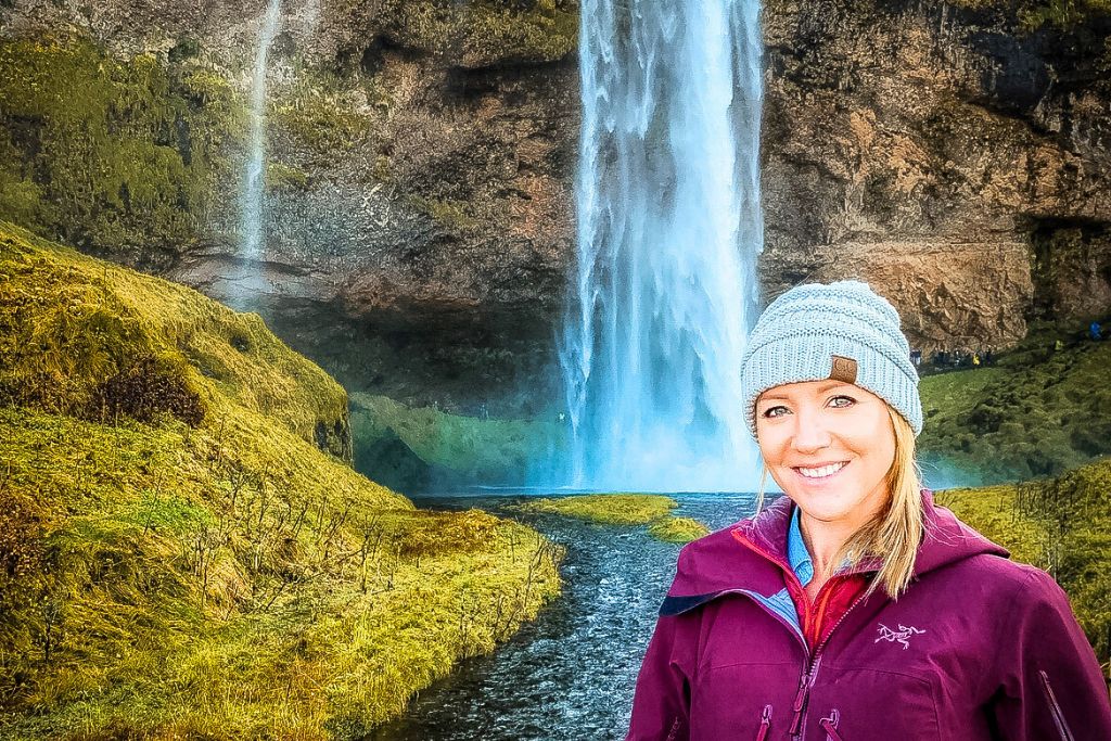 Kate smiles in front of the majestic Seljalandsfoss waterfall in Iceland. She is dressed warmly in a purple jacket and gray beanie, with the powerful waterfall cascading down behind her into a pool surrounded by lush green moss-covered cliffs. The scene captures the natural beauty and serenity of one of Iceland's iconic waterfalls.