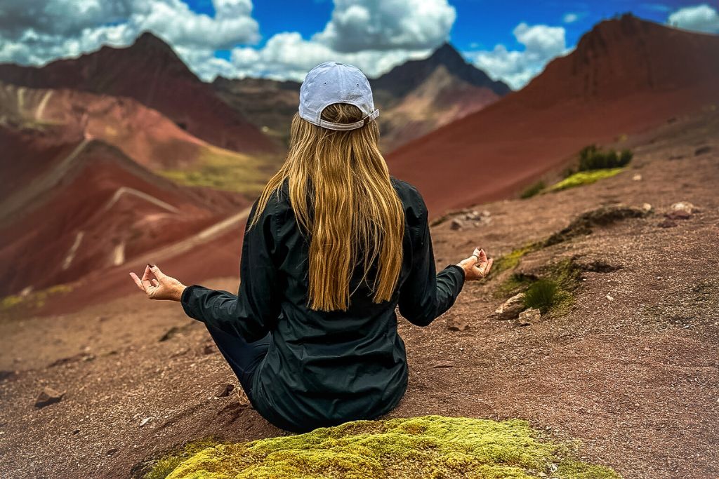 Kate is sitting cross-legged on a moss-covered rock, meditating while gazing out at the vibrant, multi-colored mountains in the background. She wears a white cap and a dark jacket, with her hands resting on her knees in a peaceful posture. The stunning landscape of red and brown earth, contrasted with a bright blue sky and clouds, creates a serene and reflective moment.