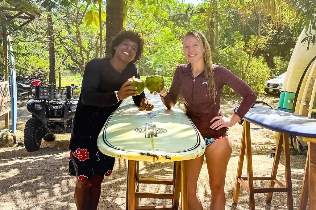 Kate and a friend stand next to surfboards while holding fresh coconuts, smiling after what appears to be a surf session. Kate wears a maroon rash guard and colorful swim shorts, while her friend sports black shorts with a red design. The backdrop features tropical trees, a parked ATV, and a laid-back beach setting, capturing a fun, relaxed vibe.
