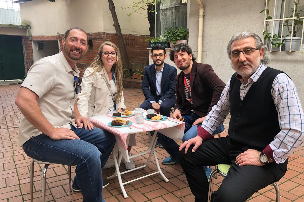 Kate and her husband sit at a small outdoor table with three locals in Istanbul, enjoying a meal to celebrate the end of Ramadan. The group is smiling and seated in a cozy courtyard, sharing traditional food and tea, creating a warm and welcoming atmosphere that highlights cultural connection and hospitality.