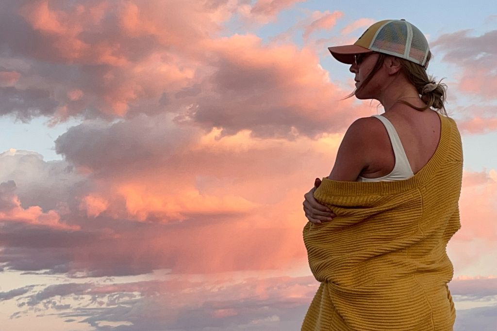 Kate stands with her arms crossed, gazing thoughtfully at a vibrant sunset sky filled with pink and orange clouds. Wearing a yellow sweater and cap, she exudes a calm and reflective energy. The serene moment embodies mindfulness, as she connects with nature in this peaceful setting.