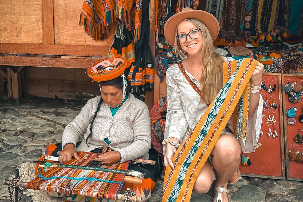 Kate sits beside a local artisan in the Sacred Valley, Peru, who is weaving intricate textiles by hand at a market. Kate smiles as she holds up a beautifully patterned, hand-woven textile, showcasing the vibrant colors and traditional craftsmanship. The scene highlights a cultural exchange and appreciation for the skill and artistry of Peruvian weaving.