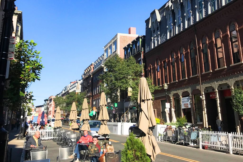 Outdoor dining along a sunny street in Boston, with tables and chairs set up beside historic red-brick buildings. Umbrellas are closed, and people are enjoying a meal, capturing the relaxed, vibrant atmosphere of a Boston neighborhood on a clear day.