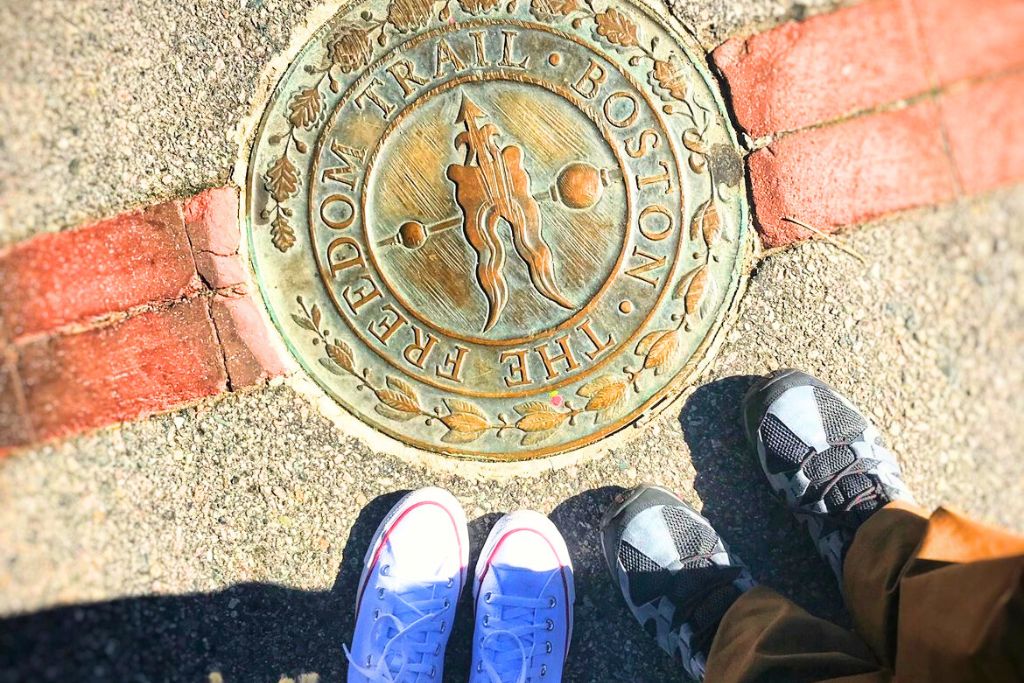 Close-up view of the Freedom Trail marker embedded in the sidewalk, a key attraction in Downtown Boston. Two pairs of shoes, one in white sneakers and the other in black, stand next to the historic marker, symbolizing a walk through Boston’s rich history.