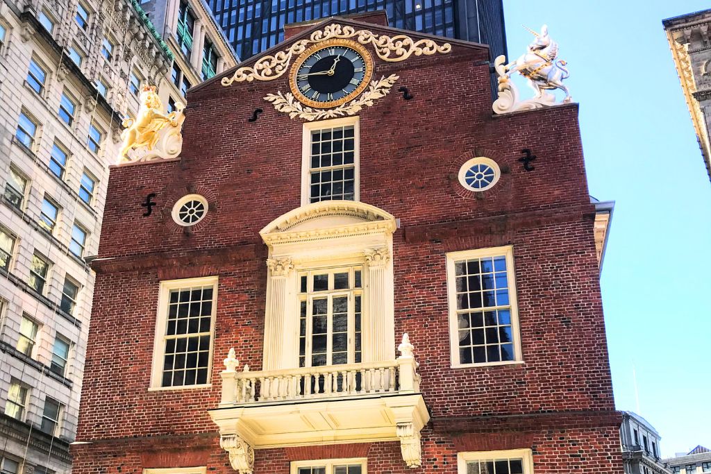 The historic Old State House in Downtown Boston, a red-brick building with a prominent clock, ornate gold decorations, and statues. Surrounded by modern skyscrapers, this landmark highlights the blend of old and new architecture in the heart of the city.