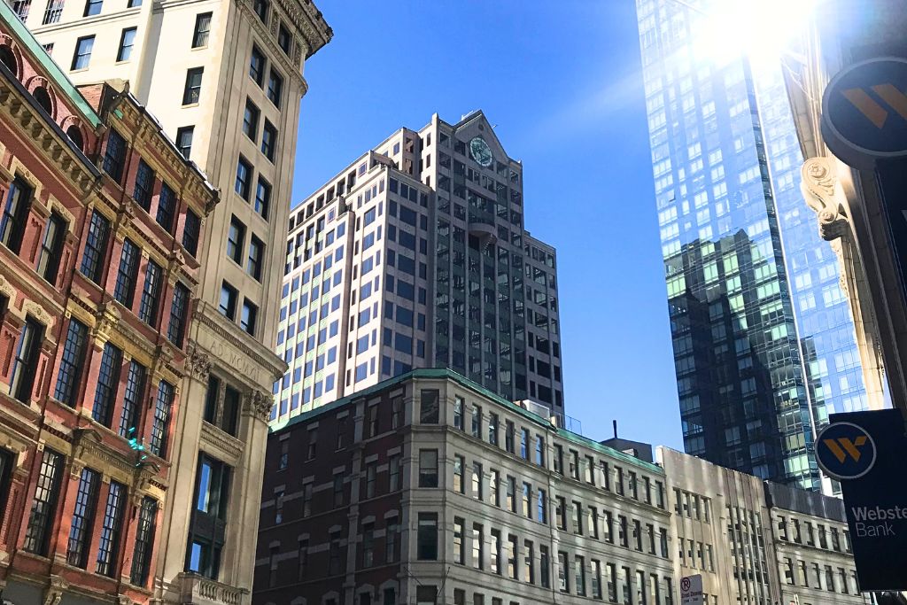 A view of tall buildings in downtown Boston under a clear blue sky, with sunlight reflecting off modern glass structures. The architecture ranges from historic red-brick facades to contemporary skyscrapers, highlighting the blend of old and new in the city.