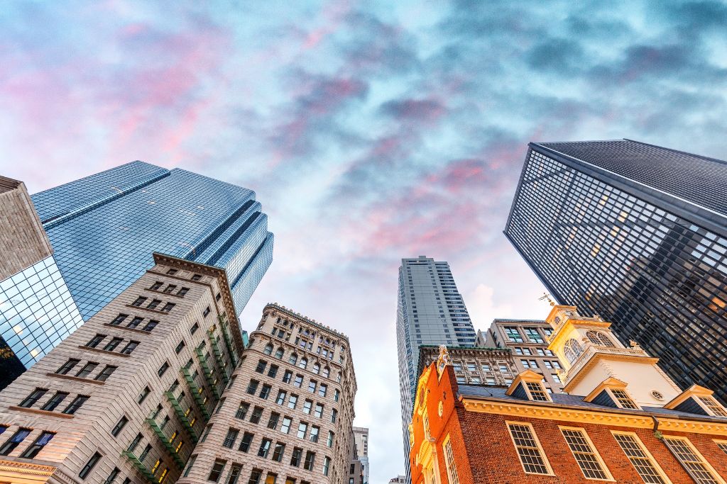 An upward view of Boston’s skyline at sunset, featuring a mix of historic and modern architecture. The Old State House, with its classic red-brick facade and clock tower, stands in contrast to the surrounding glass skyscrapers under a colorful, cloud-filled sky.