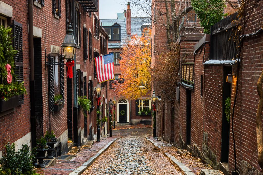 A charming cobblestone street in Beacon Hill, Boston, lined with historic red-brick buildings adorned with festive decorations and an American flag. The narrow alleyway, with its iconic gas lamps and autumnal tree in the distance, captures the quaint and historic atmosphere of this popular Boston neighborhood.