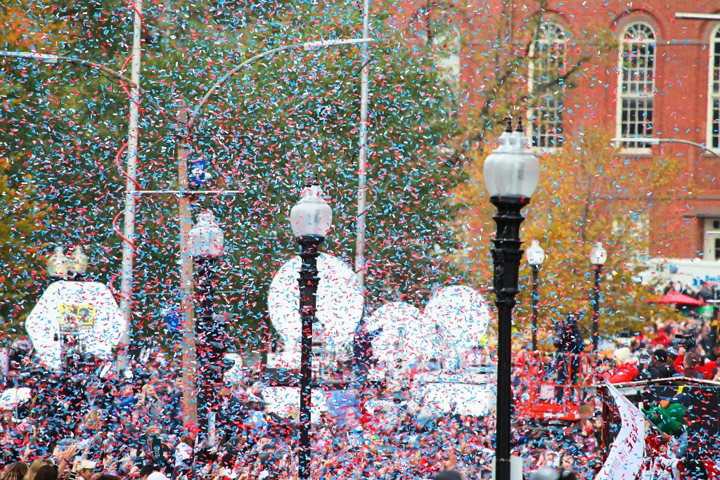 A vibrant celebration in Boston with red, white, and blue confetti filling the air above a large crowd, celebrating a Boston Red Sox victory, in front of historic brick buildings. Street lamps and fall foliage add to the lively scene, capturing the excitement of the city’s festivities.