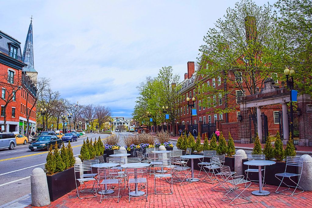 A colorful view of Harvard Square, featuring outdoor seating with metal tables and chairs on a red brick sidewalk. Historic brick buildings, tree-lined streets, and a church spire define the area, with pedestrians and cars moving through the vibrant Cambridge neighborhood near Harvard University.