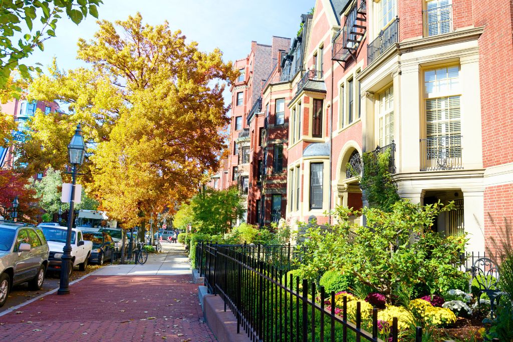 A picturesque street in Boston’s Back Bay neighborhood, featuring historic red-brick townhouses with manicured gardens and fall foliage. The tree-lined sidewalk, gas lamps, and parked cars add to the classic charm of this elegant residential area.