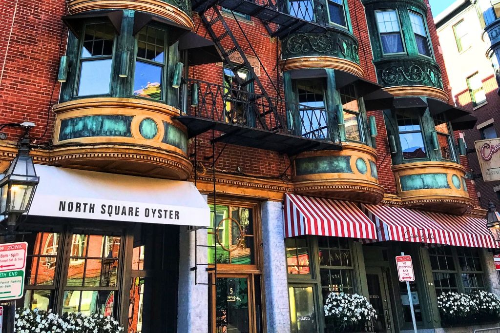 A vibrant street view of North End, Boston, featuring the historic North Square Oyster restaurant with its classic red-brick exterior, ornate bay windows, and striped awnings. The area’s charm is highlighted by the old-fashioned gas lamps and signage, reflecting the rich history of this iconic Boston neighborhood.