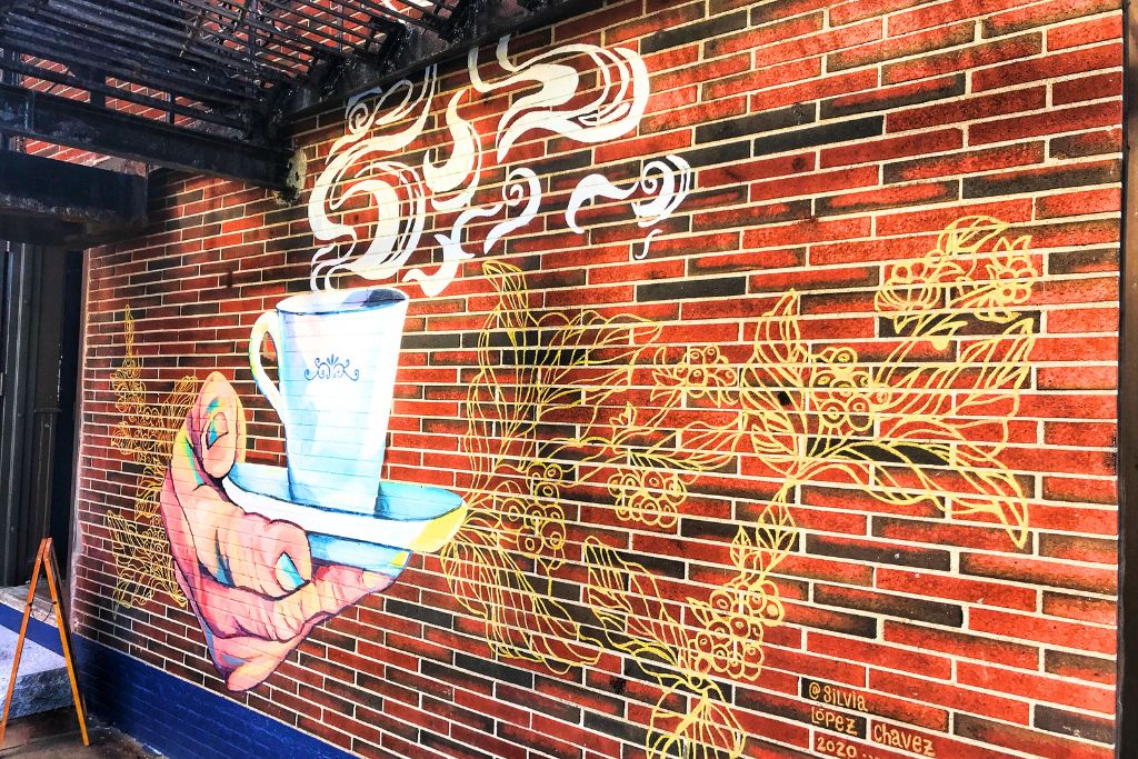 Colorful street art in Boston’s South End depicting a hand holding a steaming cup of coffee against a red brick wall. The mural, adorned with intricate gold floral designs, captures the vibrant and creative spirit of the South End neighborhood.