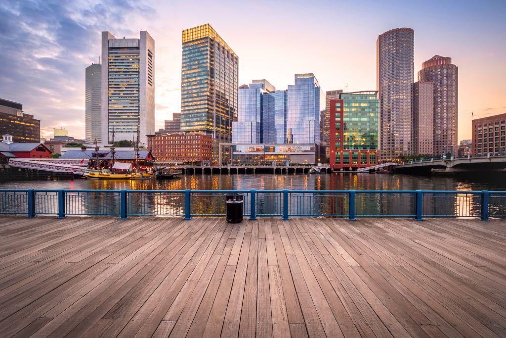 Boston waterfront at sunset, highlighting where to stay in Boston with modern skyscrapers reflecting on the water and a wooden boardwalk in the foreground. A historic sailing ship is docked along the harbor, blending the city's contemporary skyline with its maritime past.