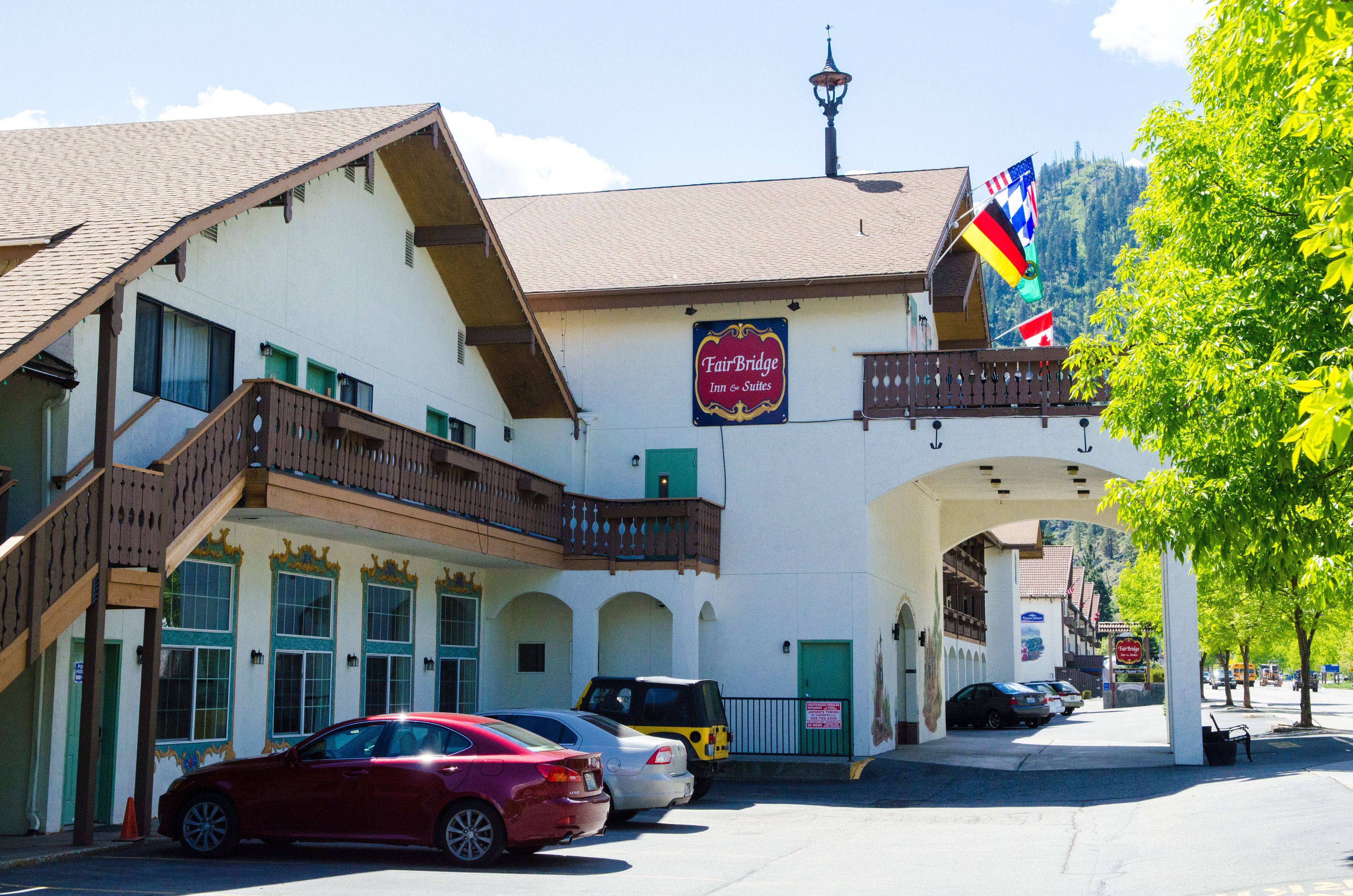 The exterior of Fairbridge Inn & Suites in Leavenworth, designed in a traditional Bavarian style with wooden balconies and decorative trims. Several flags, including the U.S., German, and Canadian flags, are displayed near the building's entrance. The structure features large windows with ornate frames and a covered walkway beneath the building. A few parked cars can be seen in front, and lush green trees frame the scene, with a backdrop of mountains in the distance.