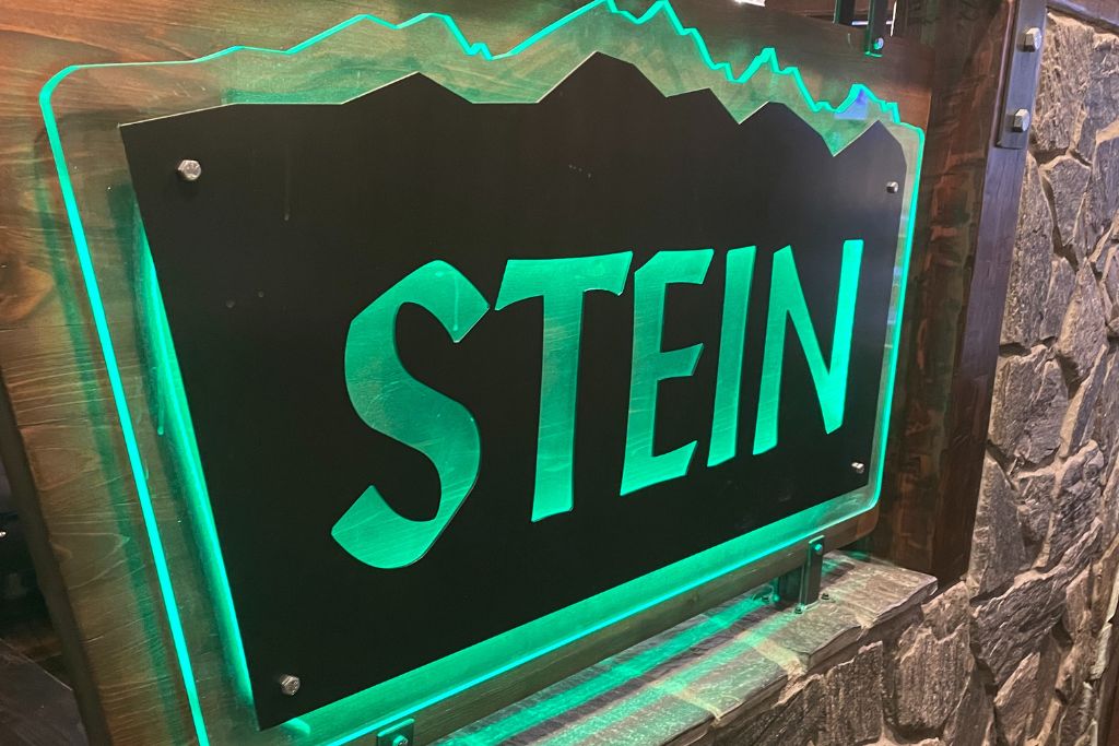 This image shows a glowing sign for "Stein," illuminated with green lighting, featuring a stylized mountain range design above the text. The sign is mounted against a stone wall, giving it a rustic yet modern feel, likely representing a German-style pub or beer hall in Leavenworth, Washington. The lighting and design suggest a warm, inviting atmosphere.