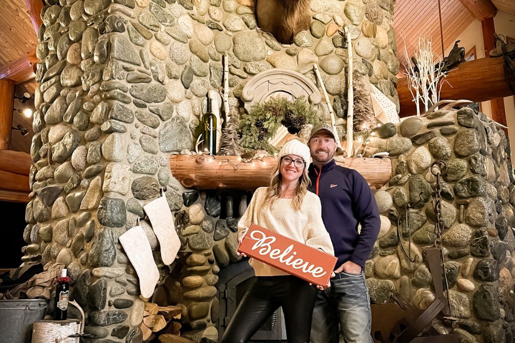 This image features Kate from Kate's Crossing Blog and her companion standing in front of a large, rustic stone fireplace at the Icicle Ridge Winery, holding a festive red sign that reads "Believe." The fireplace is decorated with stockings, greenery, and holiday-themed decor, creating a cozy and warm atmosphere. 