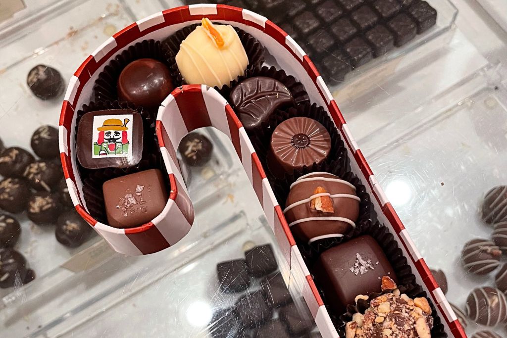 This image showcases an assortment of gourmet chocolates arranged in a candy cane-shaped box with red and white stripes. The chocolates come in various shapes and flavors, with intricate designs, including one with a festive illustration of a snowman wearing a hat. The selection includes dark, milk, and white chocolates, some topped with sea salt or nuts, creating an inviting and festive treat display.