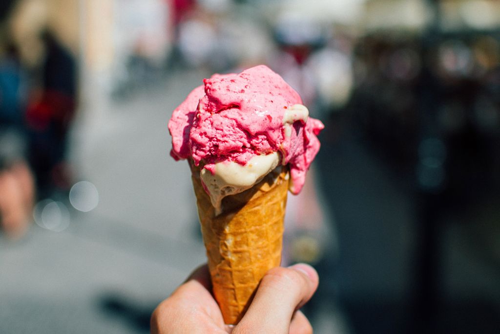 This image shows a hand holding an ice cream cone with two scoops, one of a bright pink flavor and the other of a creamy vanilla. The ice cream is slightly melting in the warm weather, creating a classic summer treat scene. The background is blurred, suggesting an outdoor location, likely a bustling street or plaza, allowing the focus to remain on the refreshing, vibrant dessert.