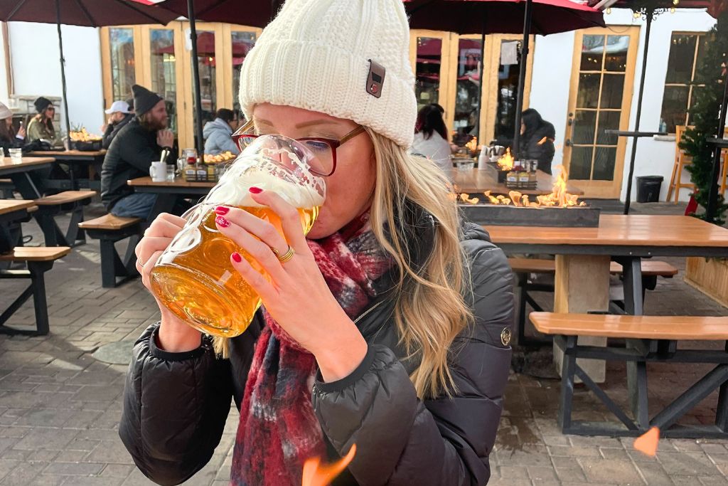 Kate from Kate's Crossing Blog is wearing a beanie and scarf, enjoying a large stein of beer at an outdoor seating area. The setting features picnic-style benches and fire pits, creating a warm, casual atmosphere despite the cold weather. Other patrons in the background are also dressed for winter, suggesting a cozy and social outdoor gathering, likely at a beer garden or pub in Leavenworth.