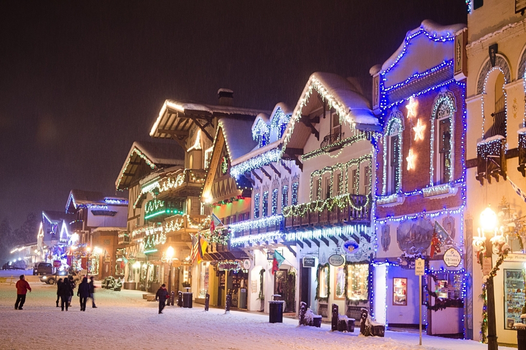 The image depicts a snowy street in Leavenworth, Washington, at night, with buildings decorated in festive holiday lights. The charming Bavarian-style architecture is highlighted by strings of colorful lights, and people are walking along the snow-covered street, adding to the wintery, holiday atmosphere. The scene captures the quaint, festive vibe of Leavenworth during the winter season.