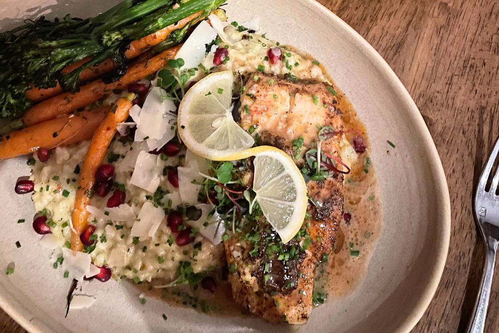 This image displays a beautifully plated dish featuring a grilled chicken breast garnished with lemon slices and fresh herbs, served on a bed of creamy risotto. The dish is accompanied by roasted baby carrots, broccolini, and scattered pomegranate seeds, adding vibrant color and texture to the meal. The presentation emphasizes a fresh, gourmet dining experience.