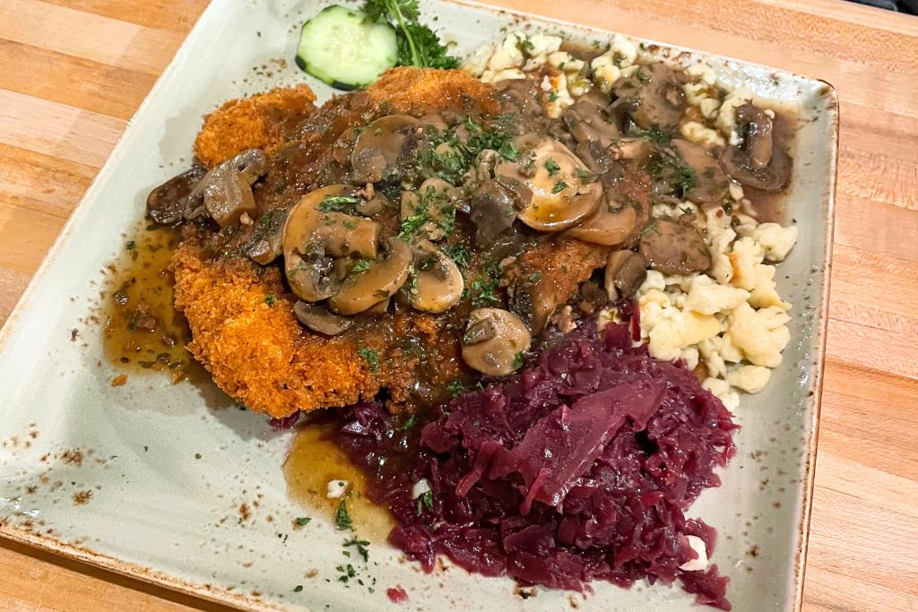 This image showcases a traditional German meal featuring a crispy breaded schnitzel topped with a rich mushroom gravy. The dish is served with a side of spätzle, a type of soft egg noodle, and tangy red cabbage. A cucumber slice and fresh parsley garnish the plate, enhancing the authentic, hearty Bavarian-style presentation.