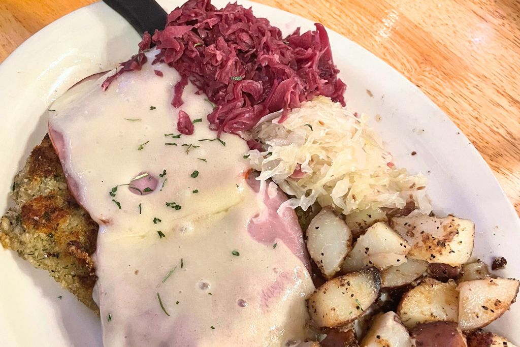 This image shows a plate featuring a traditional German-style dish with a breaded cutlet, topped with melted cheese and ham, covered in a creamy white sauce. The dish is served with sides of red cabbage, sauerkraut, and roasted potatoes, creating a hearty, comforting meal with a mix of tangy and savory flavors. The presentation reflects classic Bavarian cuisine.