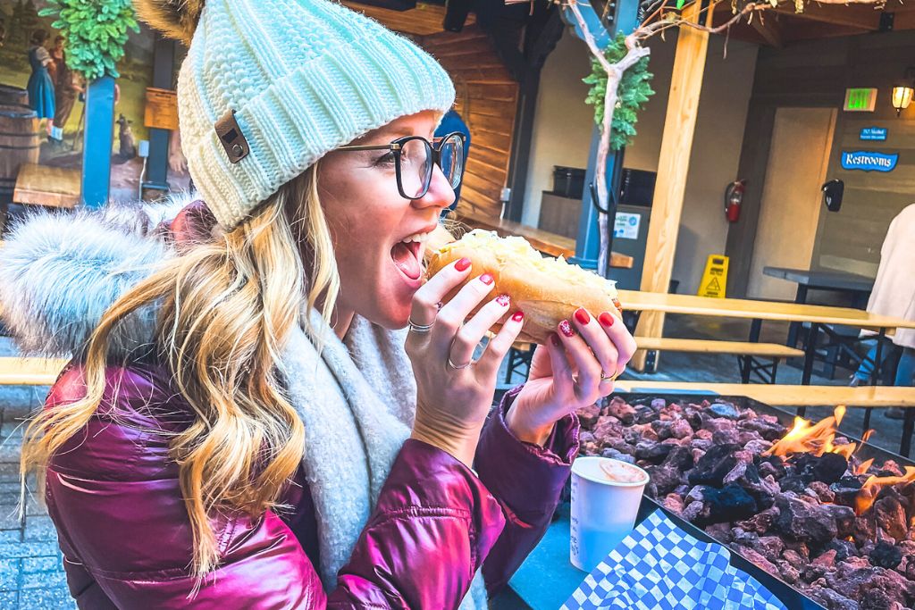 This image captures Kate from Kate's Crossing Blog enjoying a large sausage in a bun, likely a bratwurst, while sitting near an outdoor fire pit. She is dressed warmly in a beanie, scarf, and winter coat, reflecting the cold weather. The scene conveys a cozy and fun atmosphere, with the woman savoring a traditional German-style street food snack in an outdoor setting, likely in Leavenworth’s Bavarian-themed village.