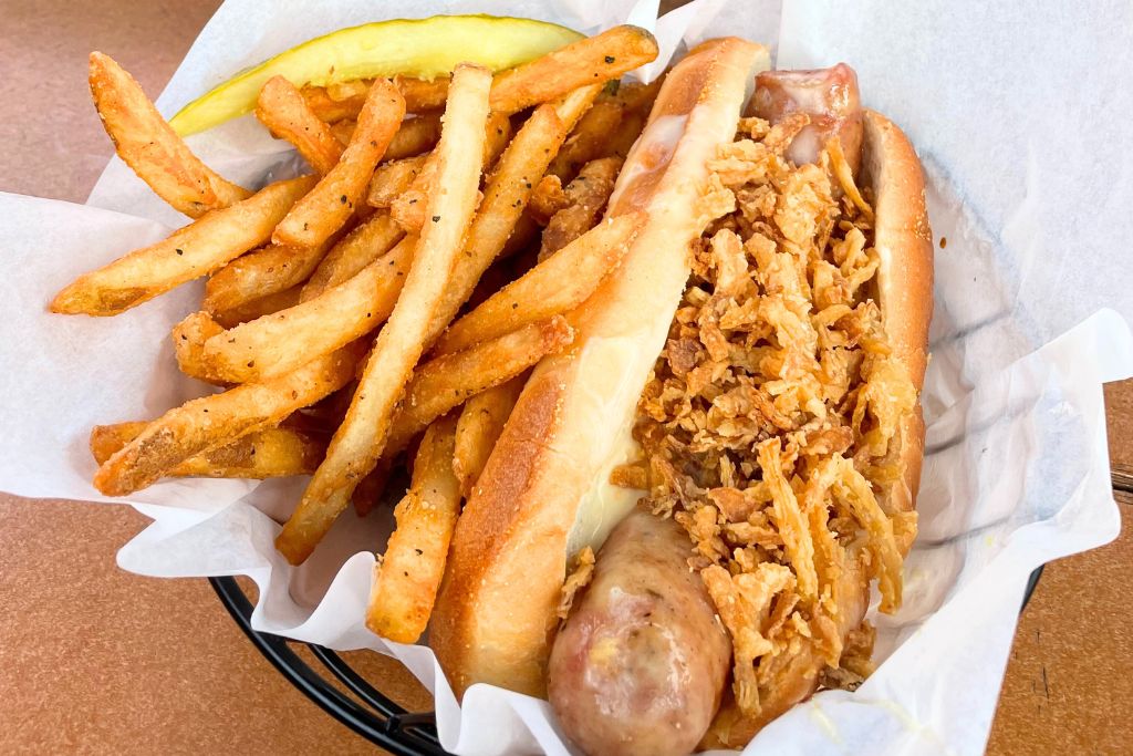 This image shows a close-up of a meal featuring a hot dog or sausage in a bun, topped with crispy fried onions and served with a side of golden French fries. A pickle spear is placed alongside the fries, adding a classic touch to the dish. The simple, satisfying combination of crispy textures and savory flavors makes for a comforting and casual meal.