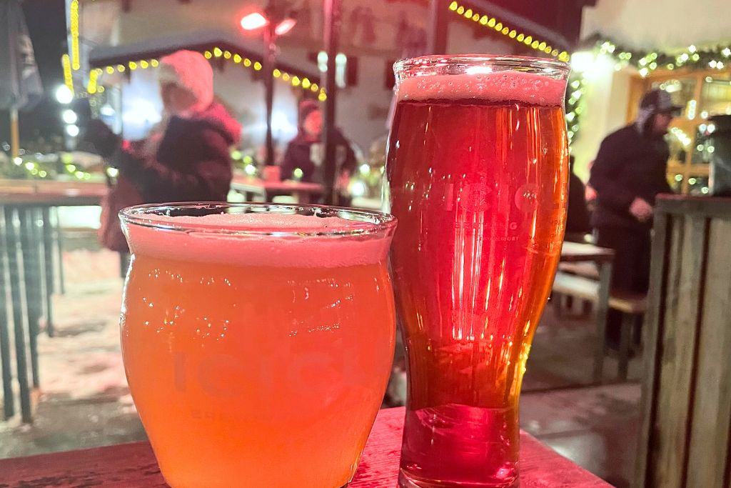 This image captures two glasses of beer, one light and one amber, set on an outdoor table in what appears to be a festive winter setting. The background shows people bundled up in warm clothing, with string lights and holiday decorations enhancing the cozy, lively atmosphere. The scene suggests an enjoyable evening at a local brewery or beer garden, possibly during a winter festival in Leavenworth.