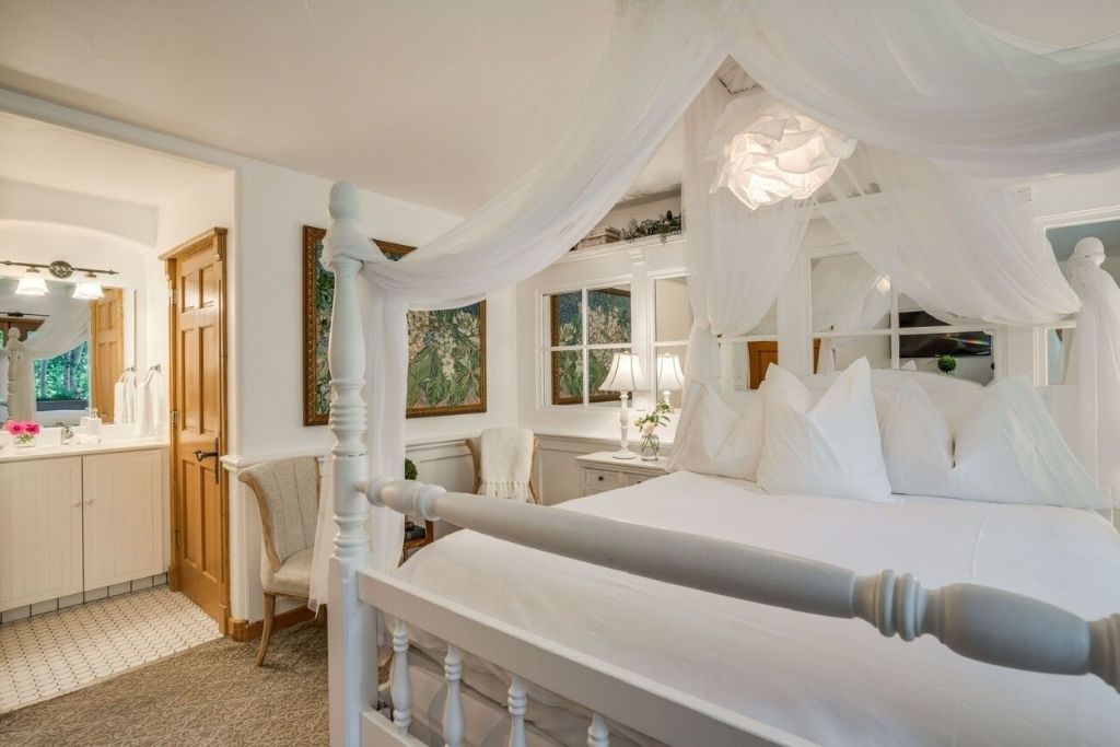 A beautifully styled room at Abendblume, featuring a luxurious canopy bed draped with delicate white fabric. The bed is adorned with plush white pillows, adding to the airy, serene ambiance. The room has soft, neutral tones and includes a cozy sitting chair and a small vanity area visible in the adjoining bathroom. Elegant decor, including framed artwork and a lamp on the bedside table, enhances the room's sophisticated charm.