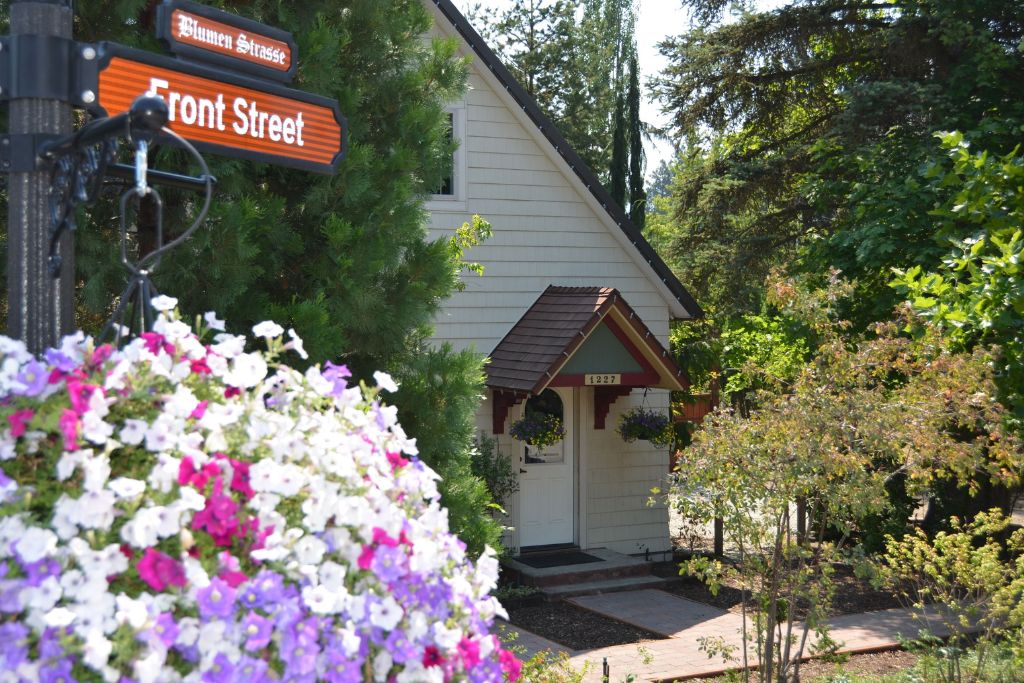 A charming cottage-style home nestled behind lush greenery on Front Street in Leavenworth. The house features a quaint entrance with hanging flower baskets and a small, gabled roof above the doorway. A sign reading "Blumen Strasse" and "Front Street" is in the foreground, adding to the picturesque, storybook-like ambiance. Bright flowers bloom in the foreground, enhancing the serene and inviting feel of this peaceful setting.