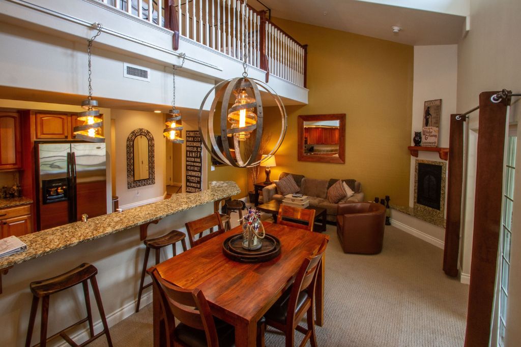 The interior of a stylish, open-concept vacation rental in Leavenworth. The space features a cozy living area with a sectional sofa, armchair, and a fireplace, creating a warm and inviting atmosphere. The dining area includes a wooden table beneath a striking chandelier, while a granite countertop with barstools separates the kitchen, which has stainless steel appliances. Above, a lofted second level overlooks the main living space, enhancing the sense of openness and light in the room.